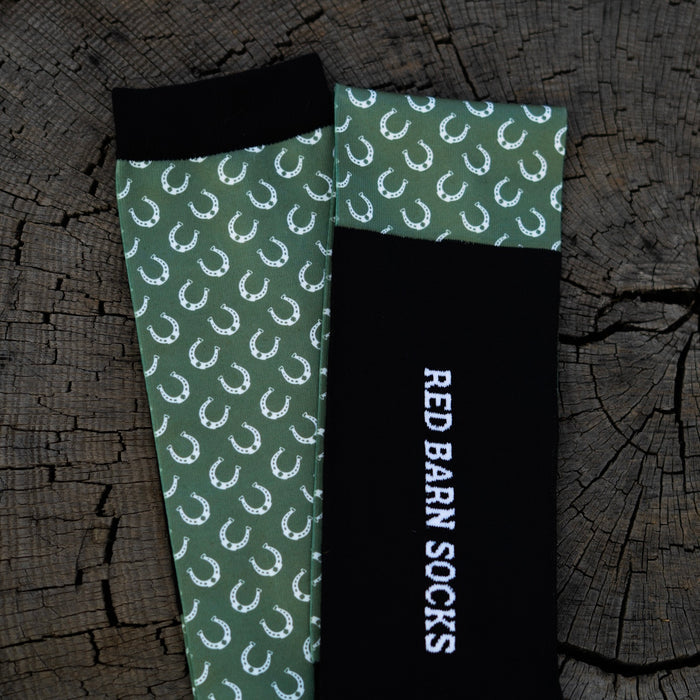 Sage green horseshoe‑pattern sock paired with a black Red Barn logo sock laid out on a tree stump