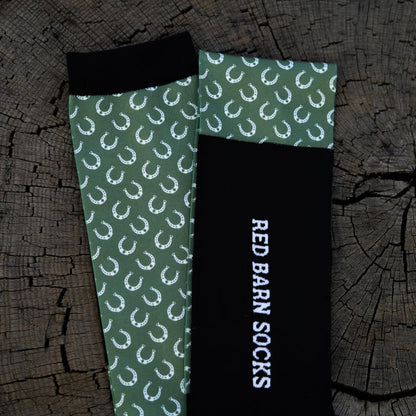 Sage green horseshoe‑pattern sock paired with a black Red Barn logo sock laid out on a tree stump