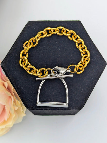 Chunky two‑tone English stirrup bracelet with large gold‑tone chain links and a silver stirrup‑style clasp displayed on a black hexagonal jewelry stand, accented by a soft pink‑and‑cream flower.